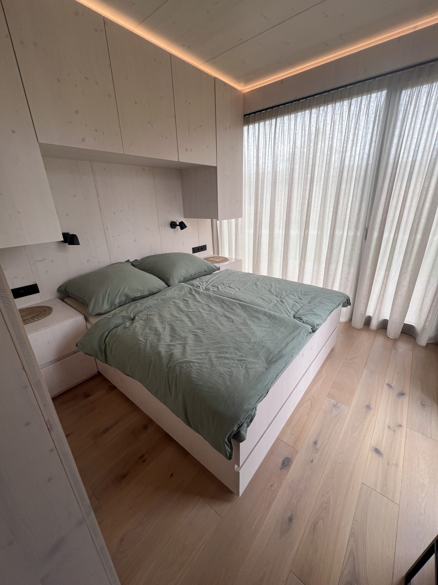 Schlafzimmer mit salbeigrüner Bettwäsche, Einbauschränken mit indirektem Licht und transparenten Vorhängen